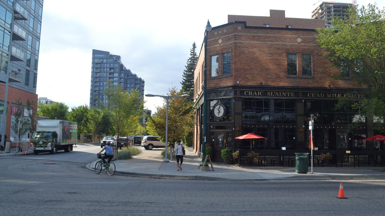 A brick corner building with a restaurant patio and umbrellas on a street in Calgary's Beltline, with towers behind, September 2017.