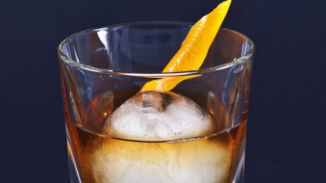 A whiskey Old Fashioned cocktail on an ice ball with an orange garnish; an illustration, not taken at Proof.