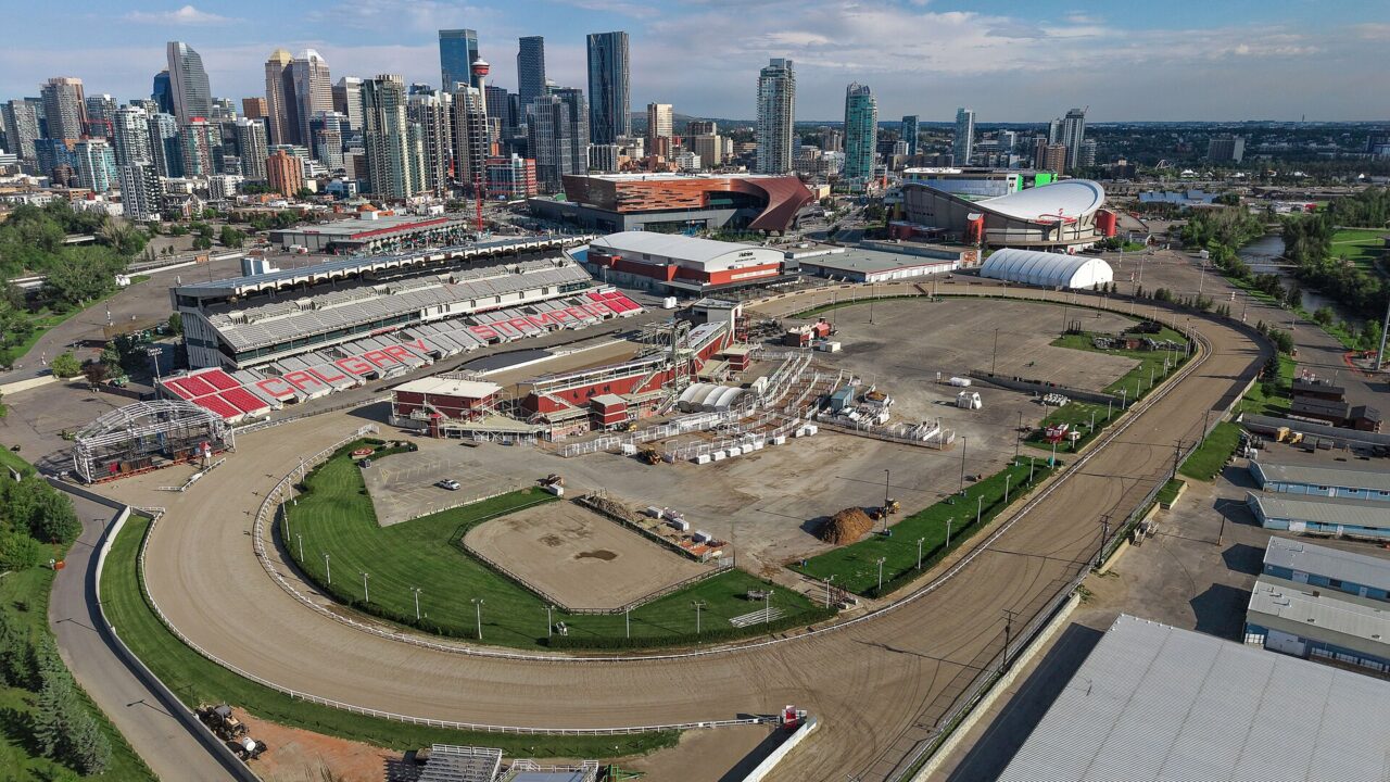 Aerial view of Stampede Park in August 2026, with the racetrack and grandstand in front, the convention centre and Saddledome behind, and the downtown Calgary skyline beyond.