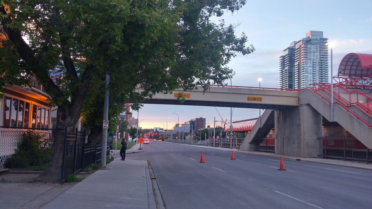 A tree-lined Victoria Park street in Calgary on a June 2017 evening, with a brick building on the left and the CTrain station footbridge ahead.
